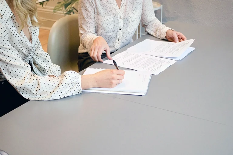 Two people in polka dot shirts sit at a desk reviewing and discussing documents, with one person pointing at the papers with a pen.