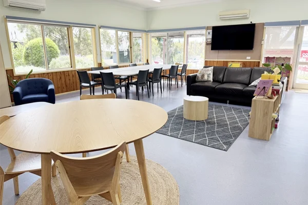 A bright, spacious room with large windows, a round wooden table with chairs, a long meeting table, black sofa, armchair, TV mounted on the wall, and shelves on a grey rug. Natural light fills the space.